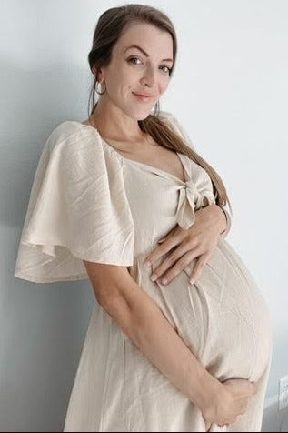 woman wearing a beige maternity dress with tie detail and flutter sleeves, cradling her baby bump