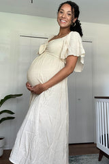 woman wearing a cream-colored maternity dress with flutter sleeves and cradling her baby bump