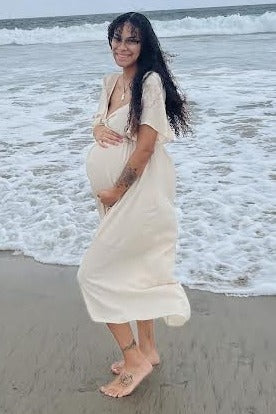 woman wearing a light beige maternity dress with short sleeves and holding her baby bump at the beach