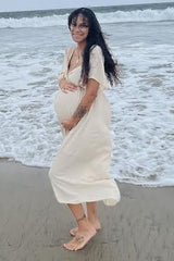 woman wearing a light beige maternity dress with short sleeves and holding her baby bump at the beach