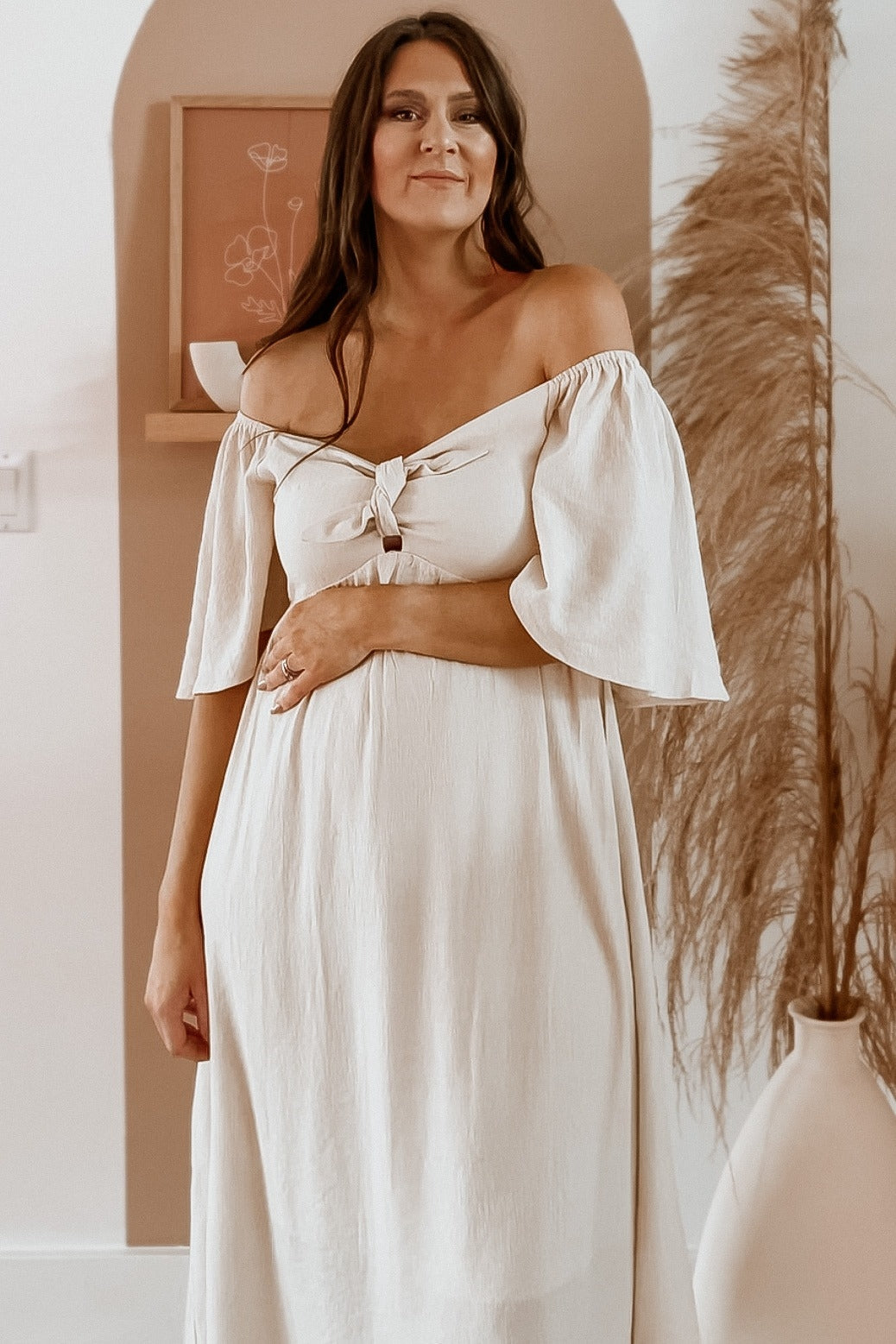 woman wearing a beige off-the-shoulder maternity dress with a tie front and flowing sleeves, holding her baby bump