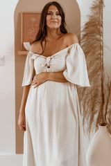 woman wearing a light beige off-the-shoulder maternity dress with a front tie and short flared sleeves, holding her baby bump