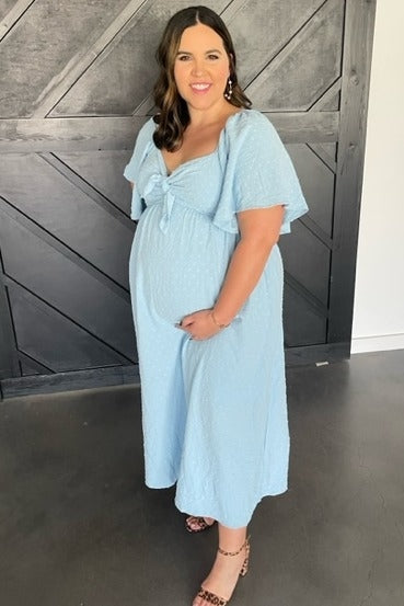 woman wearing a light blue maternity dress with short flutter sleeves and a knotted bodice, cradling her baby bump