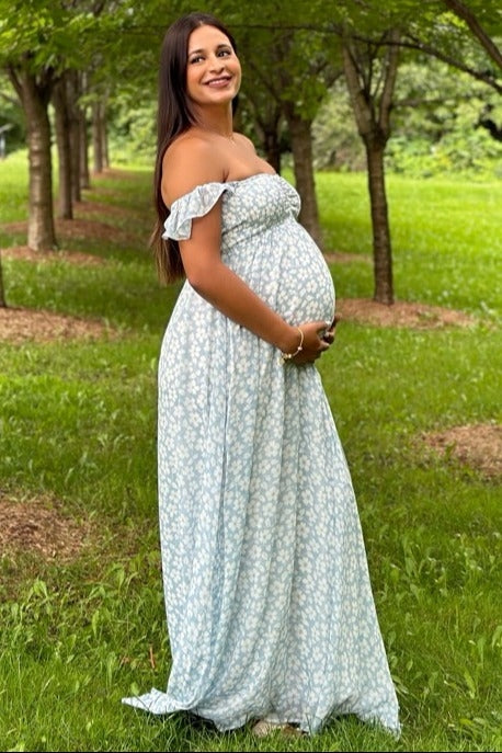 woman wearing a light blue off-shoulder maternity maxi dress and holding her baby bump in a green park