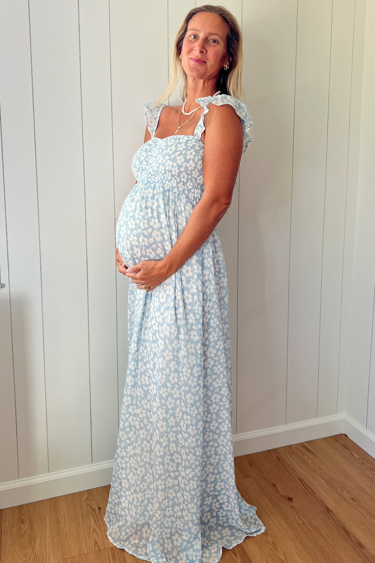 woman wearing a light blue maternity maxi dress with floral patterns and ruffle straps, holding her baby bump