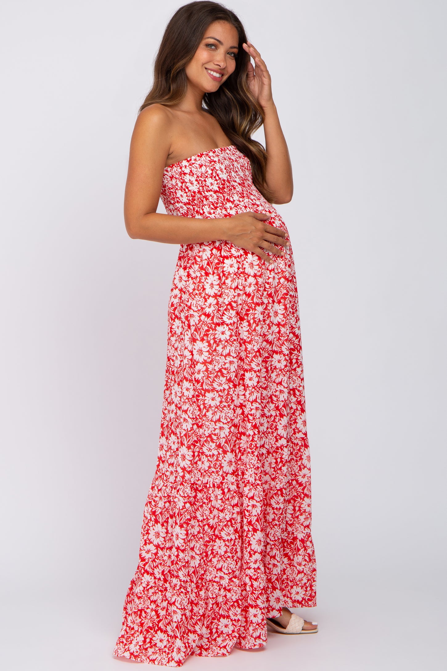 woman wearing a red and white floral strapless maternity maxi dress and holding her baby bump