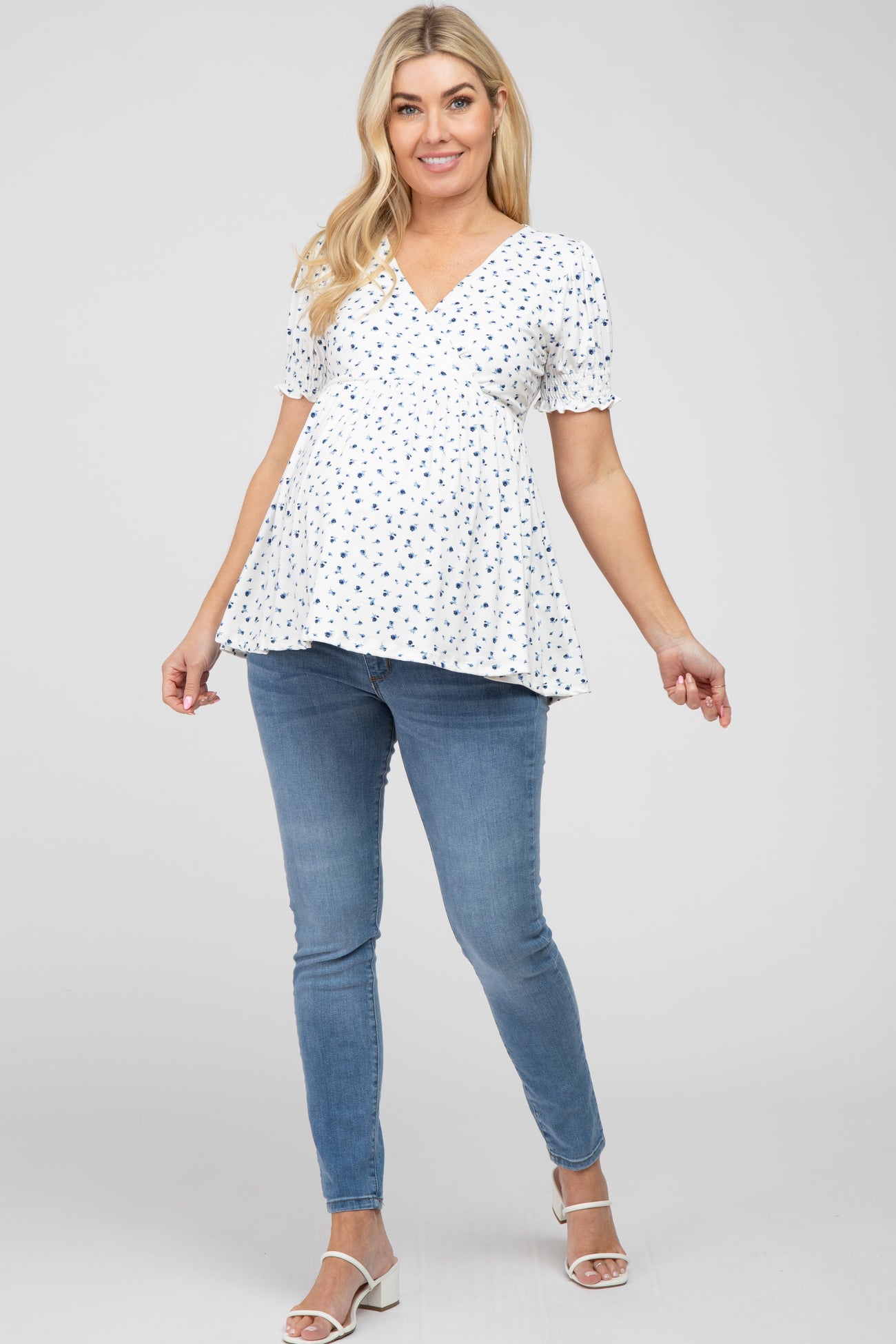 pregnant woman wearing a white floral maternity top and blue skinny jeans with white heels