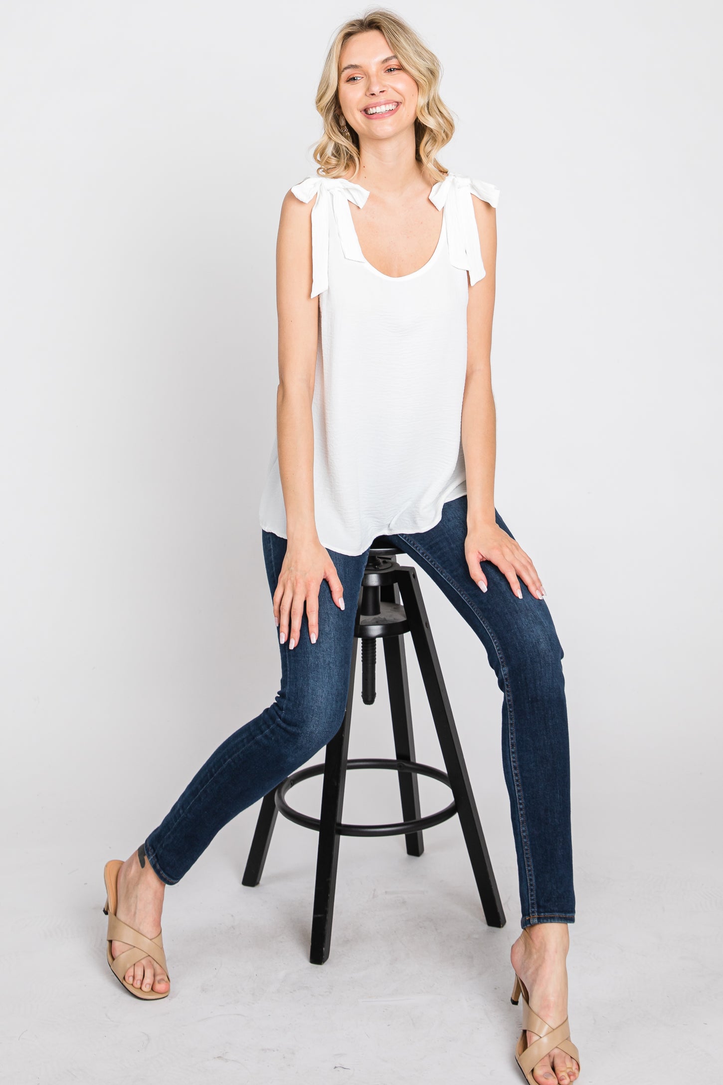 woman wearing a white sleeveless top with bow tie details, paired with dark skinny jeans, sitting on a stool and smiling
