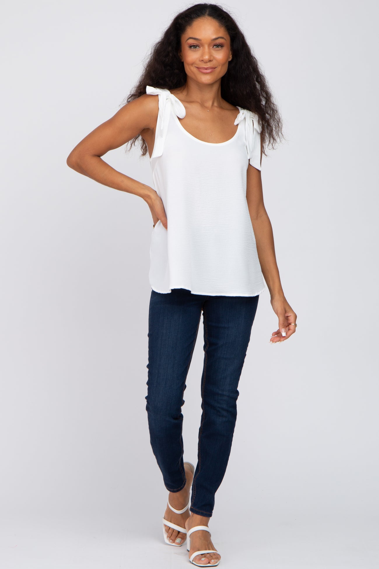 woman wearing a white sleeveless top with tie straps and blue skinny jeans, posing with one hand on her hip