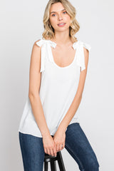 woman wearing a white sleeveless top with tie details on the shoulders and sitting on a stool