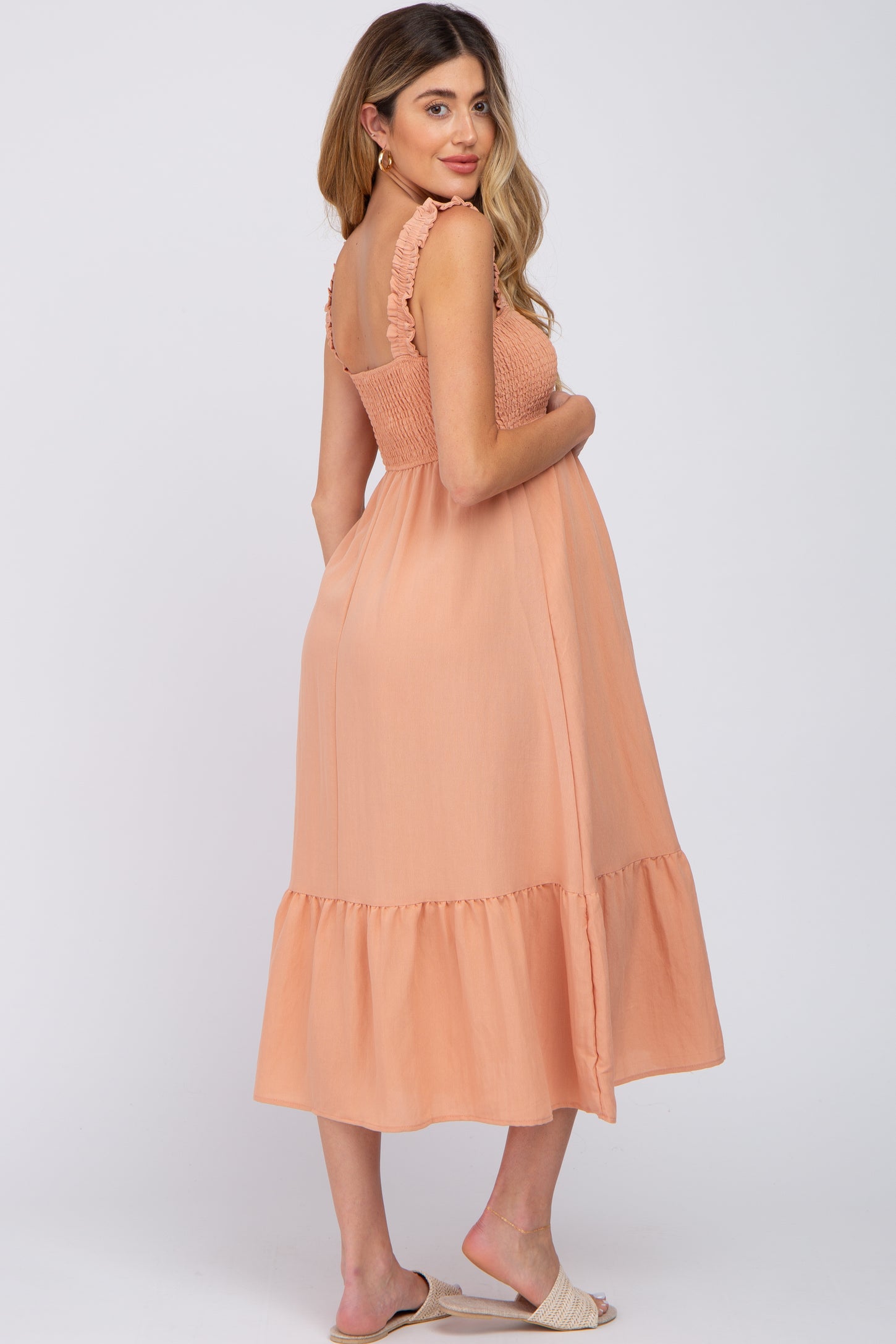 woman wearing a peach maternity dress with ruffled straps and a tiered hem, gently cradling her baby bump