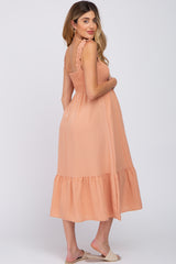 woman wearing a peach maternity dress with ruffled straps and a tiered hem, gently cradling her baby bump