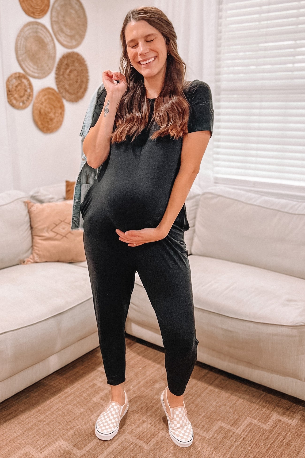 woman wearing a black maternity jumpsuit, showcasing her baby bump, standing in a cozy living room setting