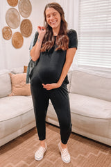 woman wearing a black maternity jumpsuit, showcasing her baby bump, standing in a cozy living room setting