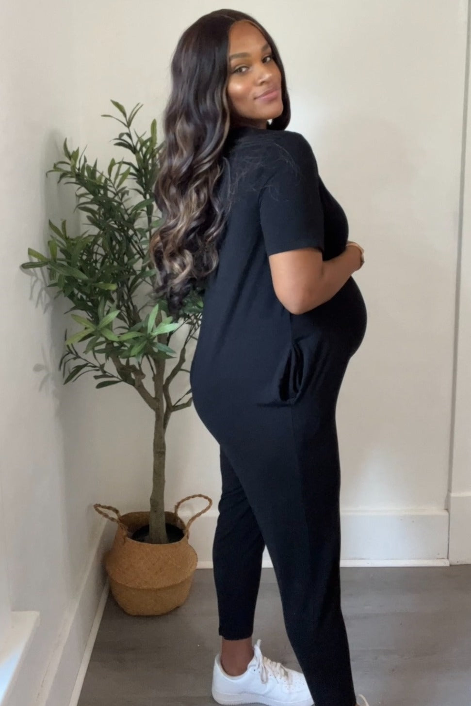 woman wearing a black maternity jumpsuit and showing her baby bump while posing next to a potted plant
