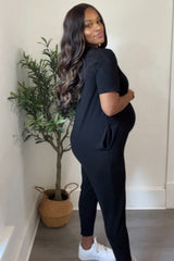 woman wearing a black maternity jumpsuit and showing her baby bump while posing next to a potted plant