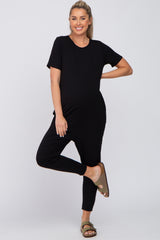 woman wearing a black short sleeve maternity jumpsuit and posing with one leg lifted, showcasing her baby bump