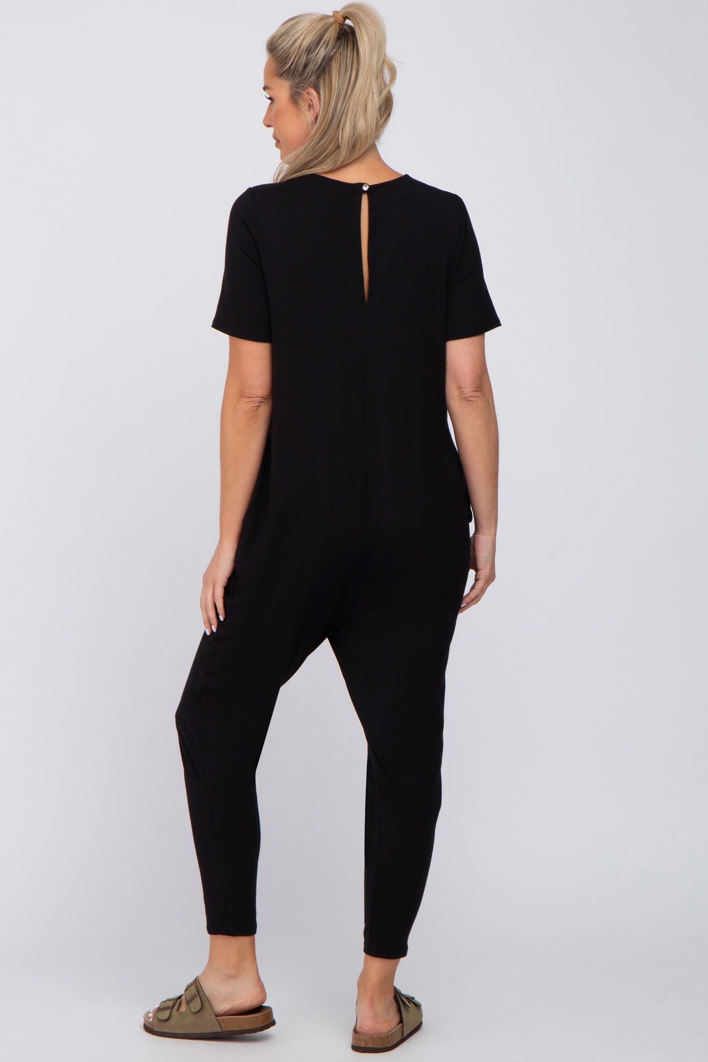 Back view of a woman wearing a black short sleeve maternity jumpsuit with a keyhole back and olive green sandals