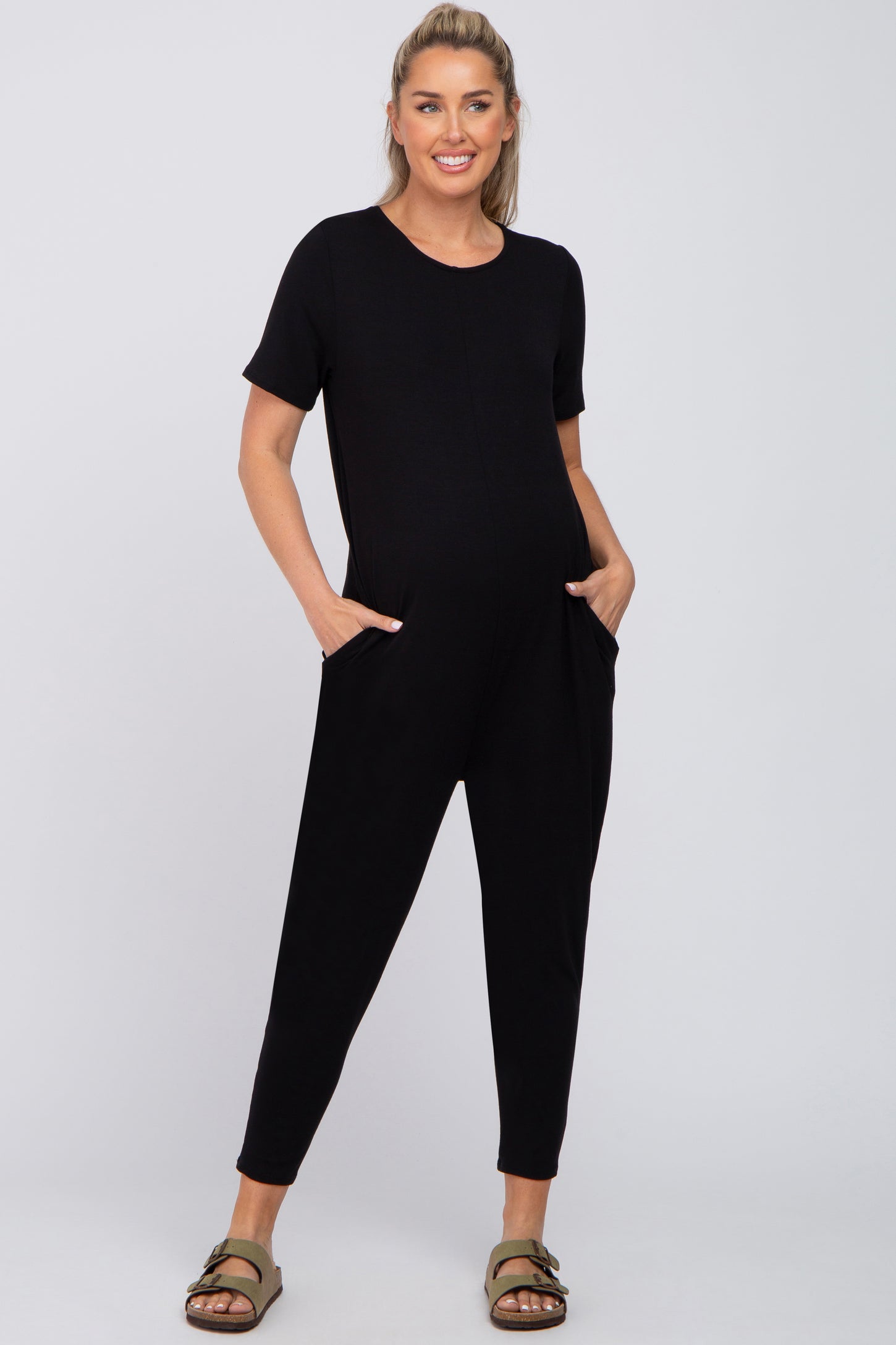 pregnant woman wearing a black short sleeve maternity jumpsuit with pockets and olive green sandals