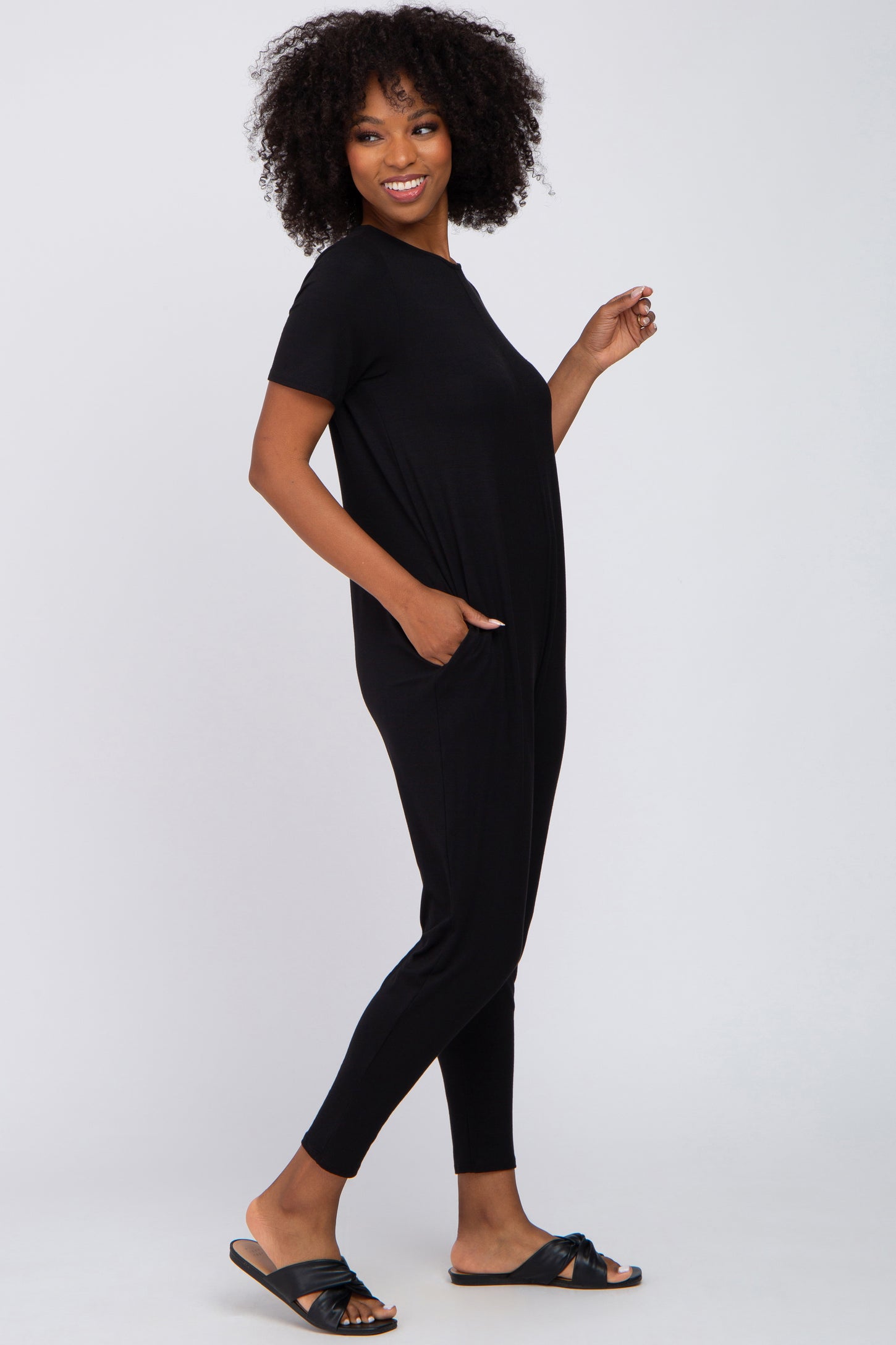 woman wearing a black short sleeve jumpsuit with pockets, standing sideways with curly hair and flat sandals