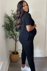 woman wearing a black maternity jumpsuit and showing her baby bump while standing next to a plant
