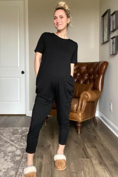 woman wearing a black maternity jumpsuit with side pockets and a relaxed fit, standing in a cozy living room
