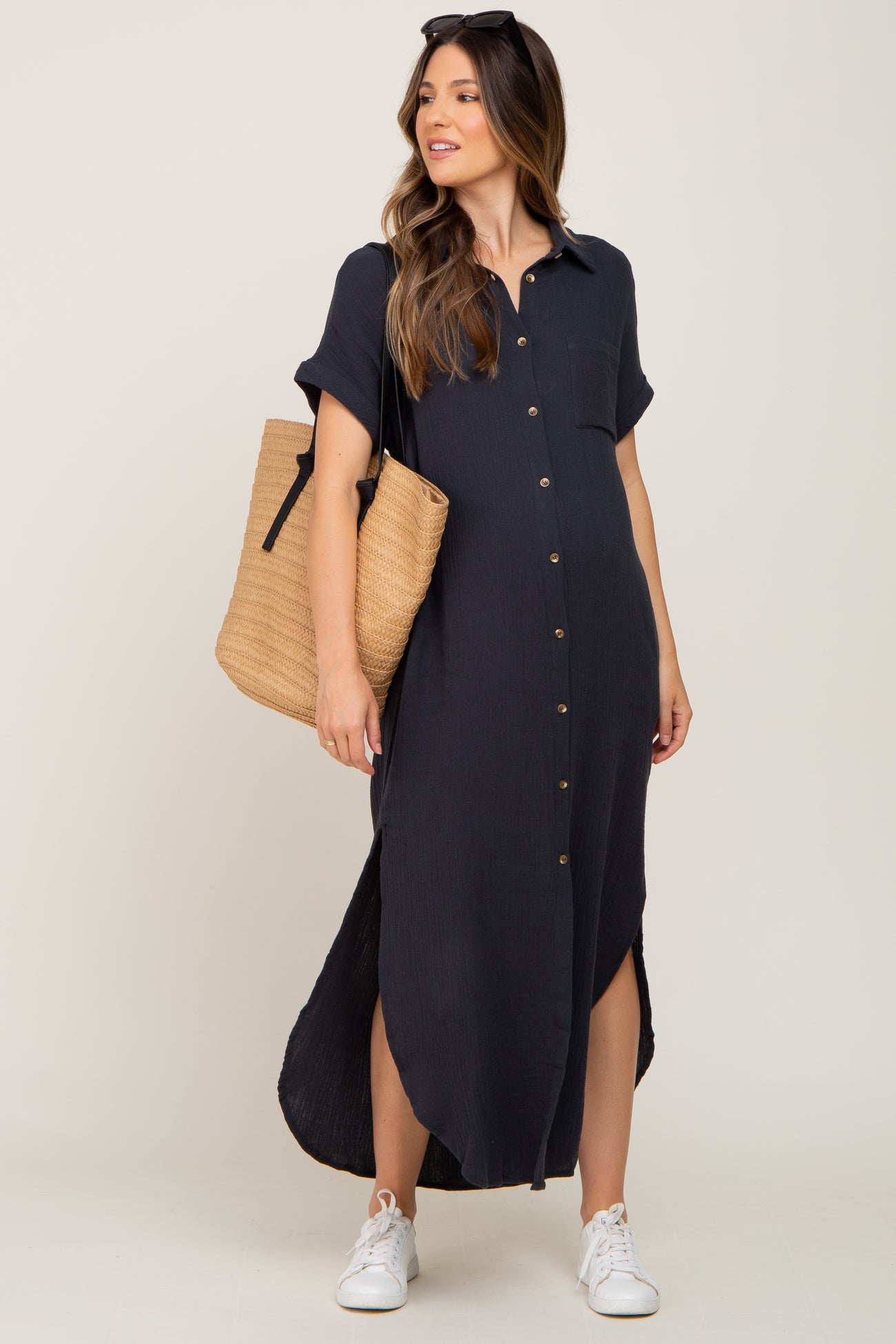 woman wearing a navy blue short sleeve button-up maternity dress with side slits, holding a straw tote bag