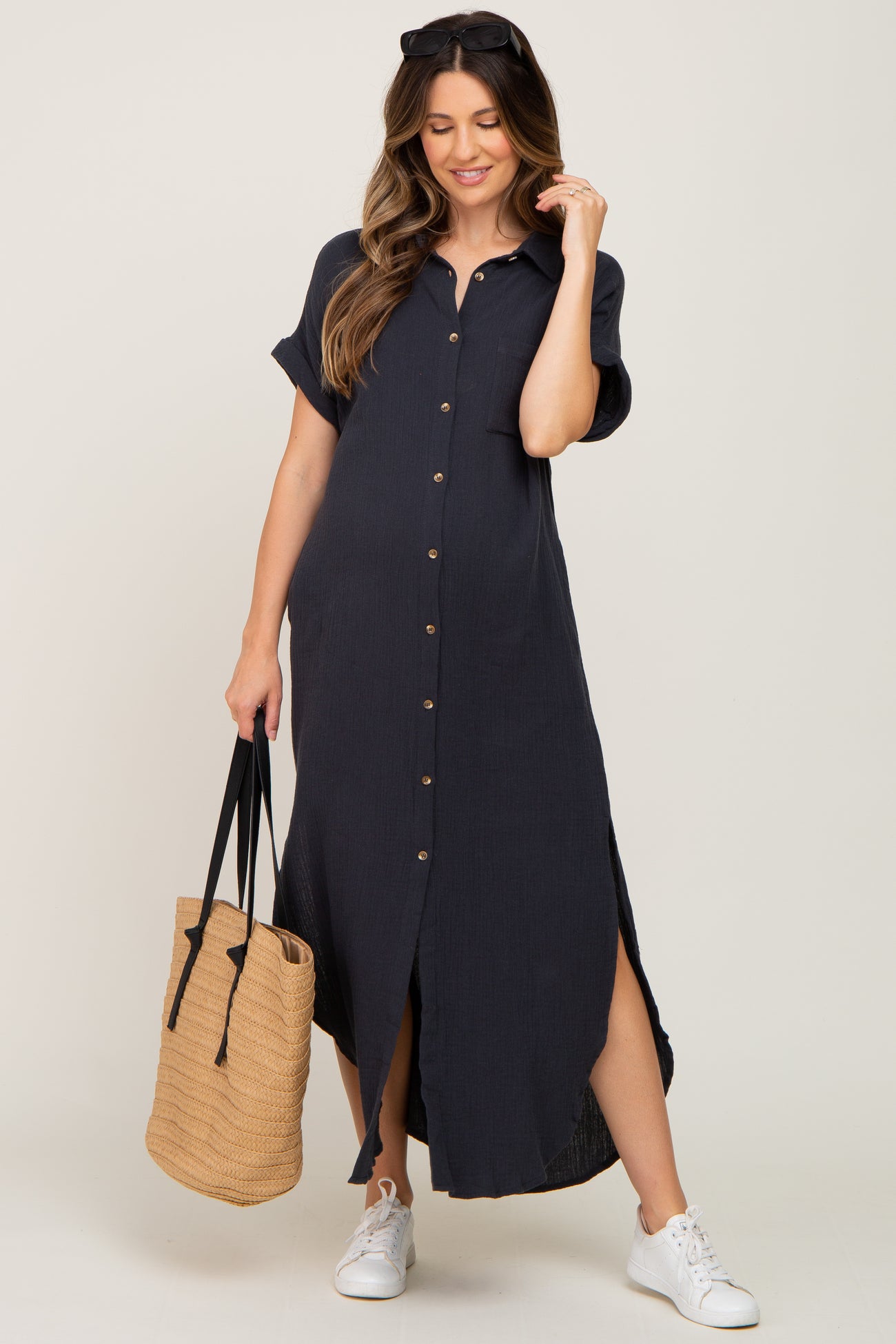 woman wearing a navy blue short sleeve button-up maternity dress with side slits, holding a woven bag and wearing sneakers