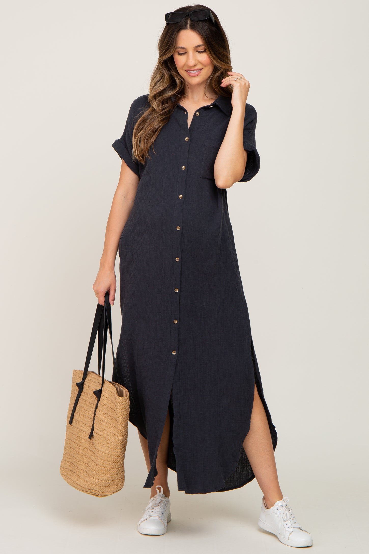 woman wearing a navy blue short sleeve button-up maternity dress with side slits, holding a woven bag and wearing sneakers