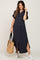 woman wearing a navy blue short sleeve button-up maternity dress with side slits, holding a woven bag and wearing sneakers