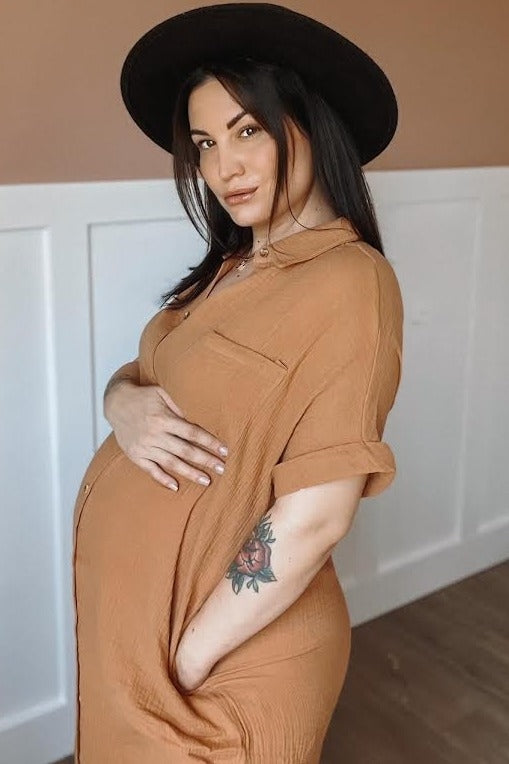 woman wearing a rust-colored maternity dress with a front pocket, showcasing her baby bump and wearing a black hat