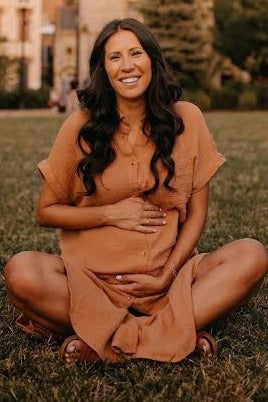 woman wearing a rust-colored short sleeve maternity dress, sitting on grass and holding her baby bump