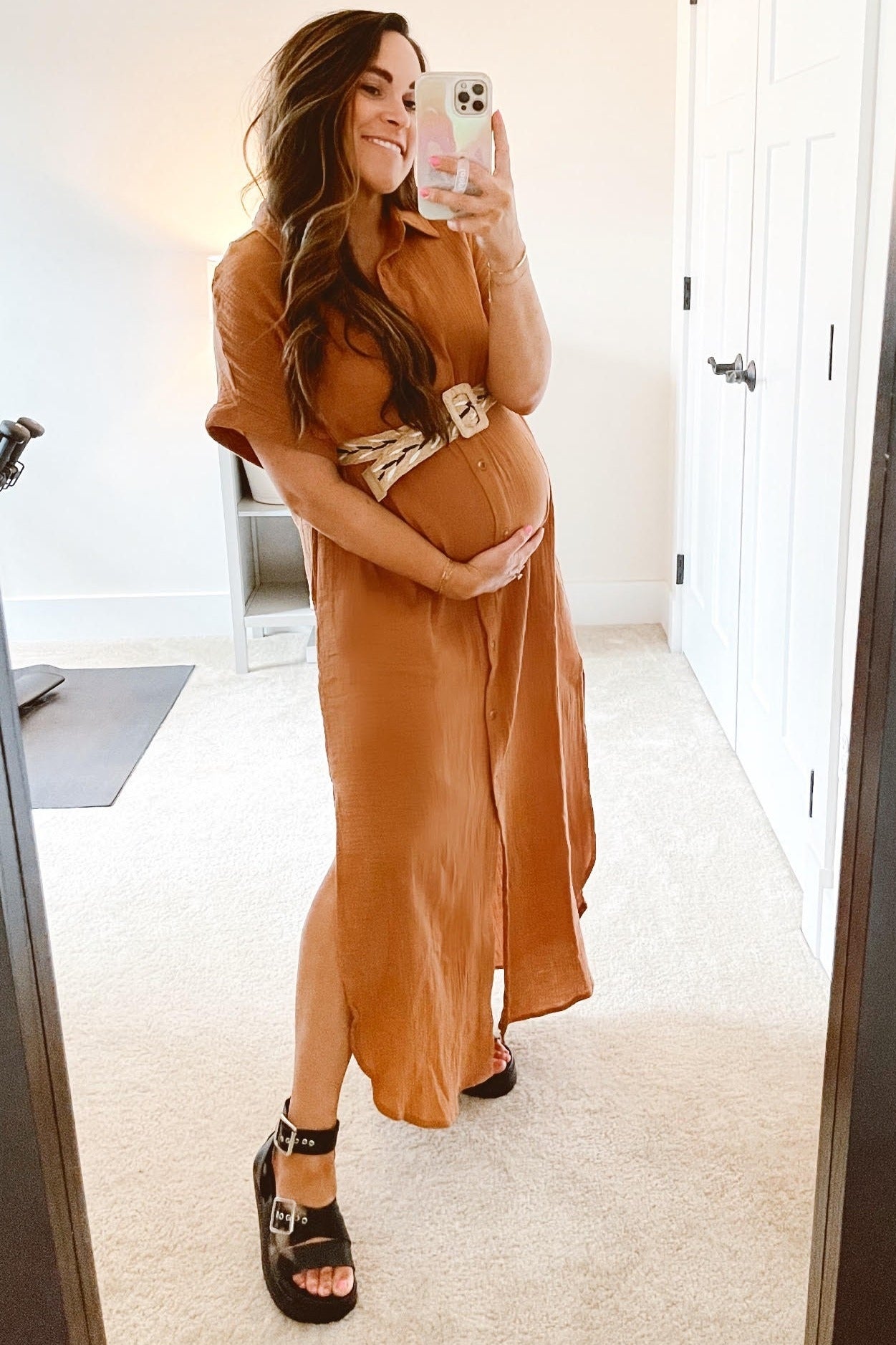 woman wearing a rust-colored short sleeve maternity dress with a belted waist and holding her baby bump in front of a mirror