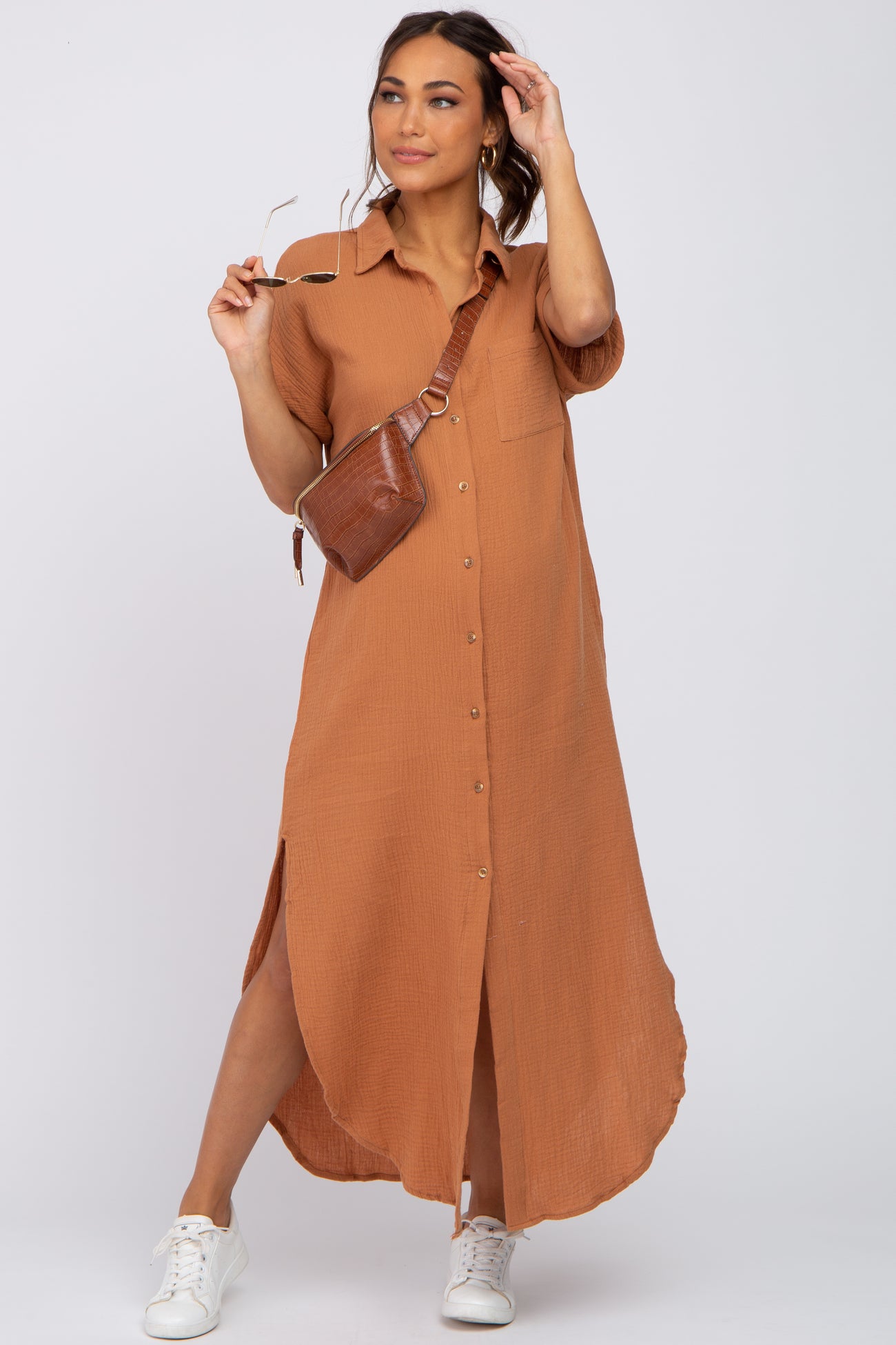woman wearing a brown maternity shirt dress with buttons and side slits, styled with white sneakers and a brown crossbody bag