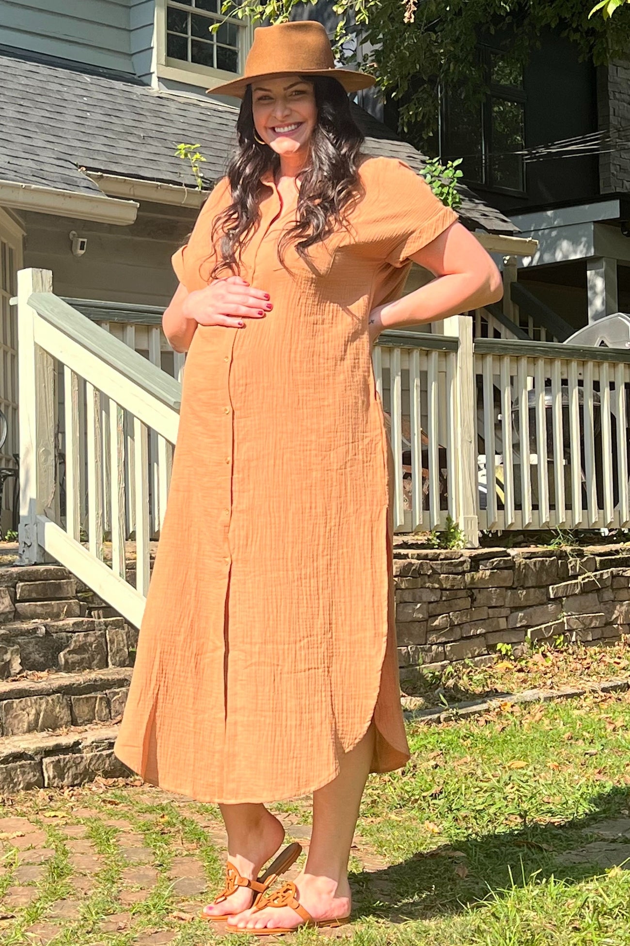 woman wearing a rust-colored maternity dress with buttons and holding her baby bump outdoors