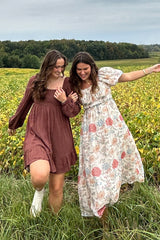 Two women in dresses laughing and walking through a yellow flower field with trees in the background