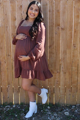 Smiling pregnant woman in a mauve maternity dress and white boots posing against a wooden fence