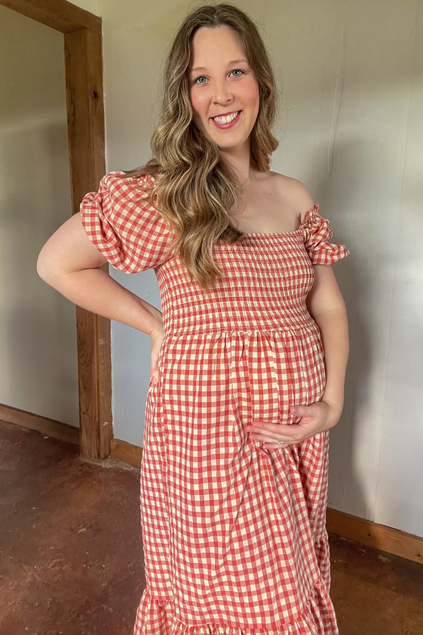 woman wearing a red and white checkered off-the-shoulder maternity dress and cradling her baby bump