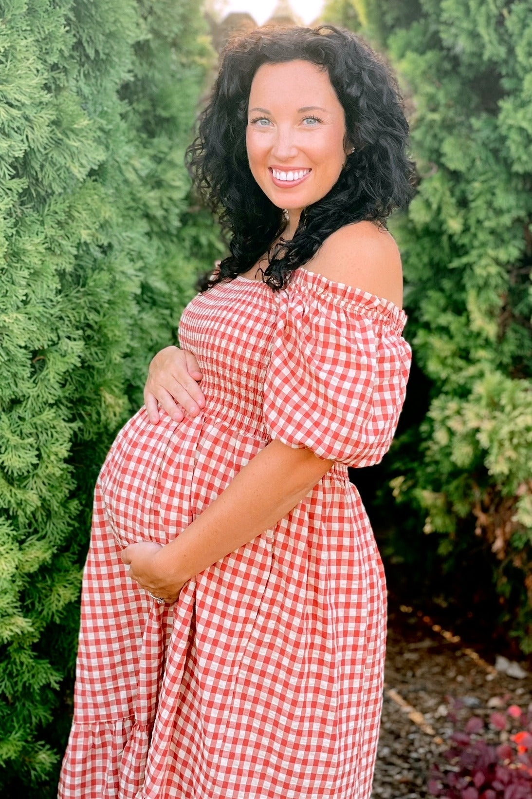 woman wearing a red and white checkered off-the-shoulder maternity dress and holding her baby bump