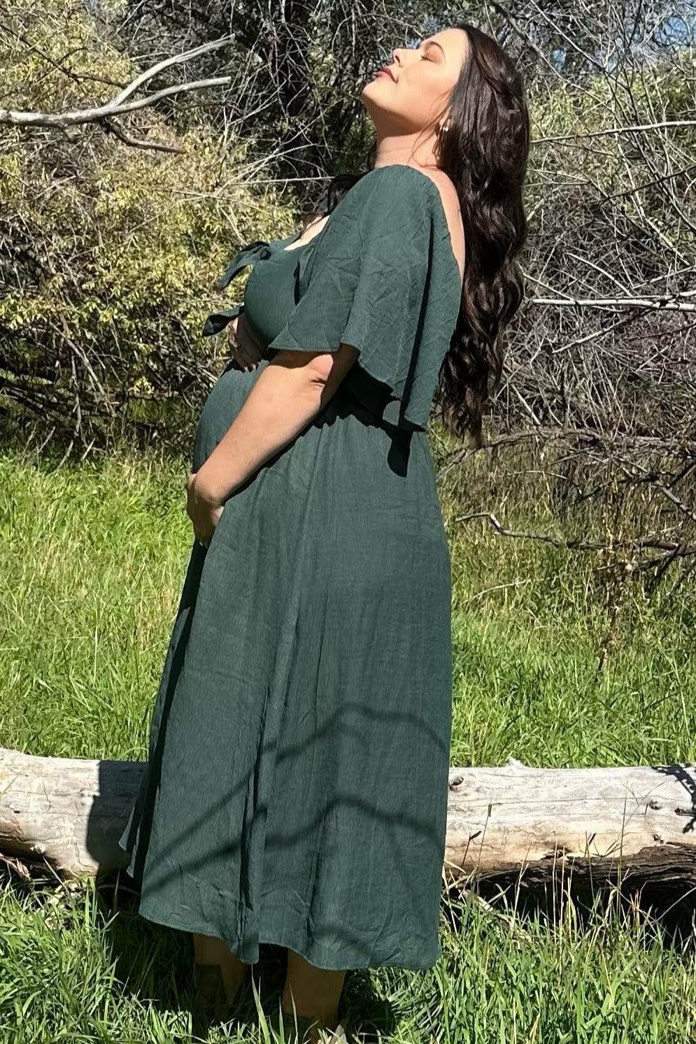 woman wearing a dark green maternity dress with flutter sleeves and holding her baby bump in an outdoor setting