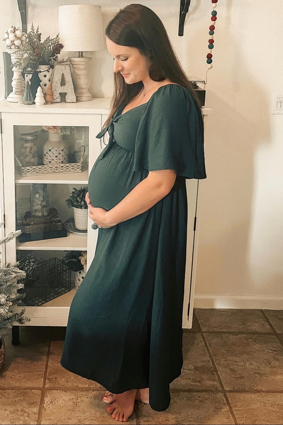 woman wearing a dark green maternity maxi dress and showing her baby bump while standing indoors