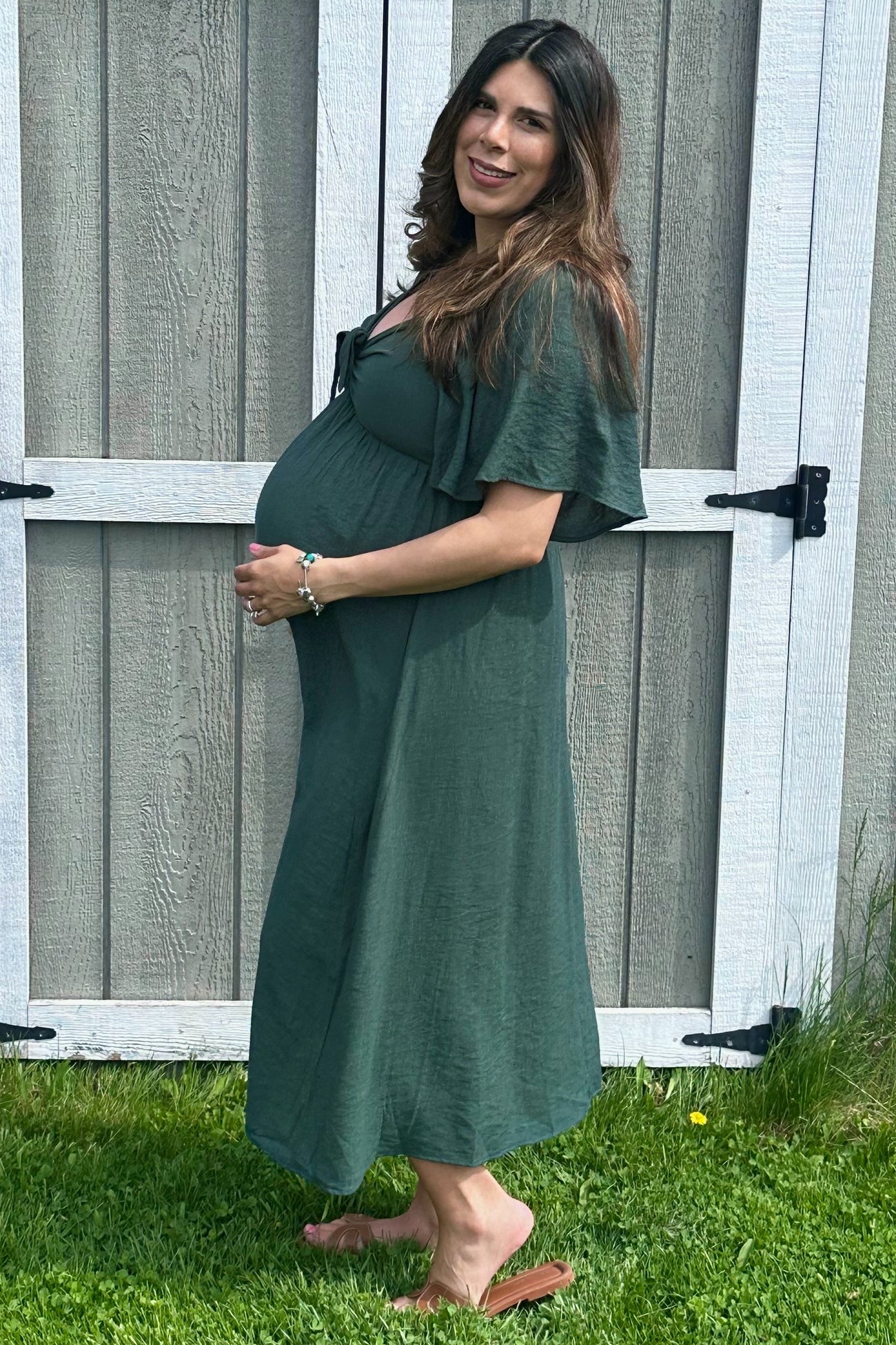 woman wearing a dark green maternity dress with flutter sleeves and holding her baby bump outdoors