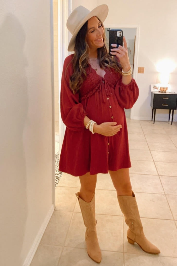 woman wearing a burgundy maternity mini dress with buttons and bell sleeves, holding her baby bump, paired with tan knee-high boots and a wide-brimmed hat