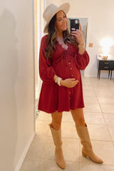 woman wearing a burgundy maternity mini dress with buttons and bell sleeves, holding her baby bump, paired with tan knee-high boots and a wide-brimmed hat