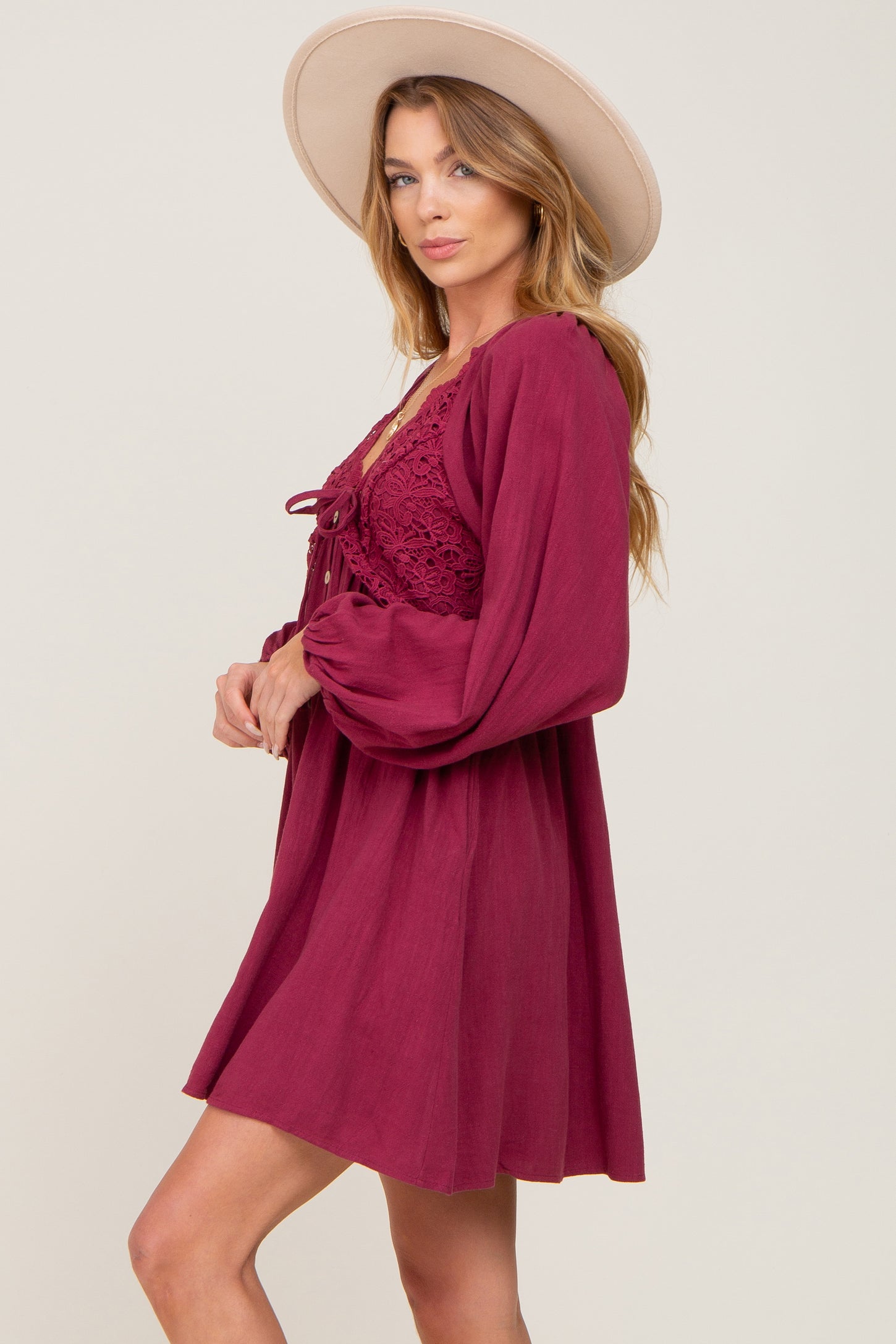 woman wearing a burgundy short dress with lace details and long sleeves, accessorized with a wide-brimmed hat