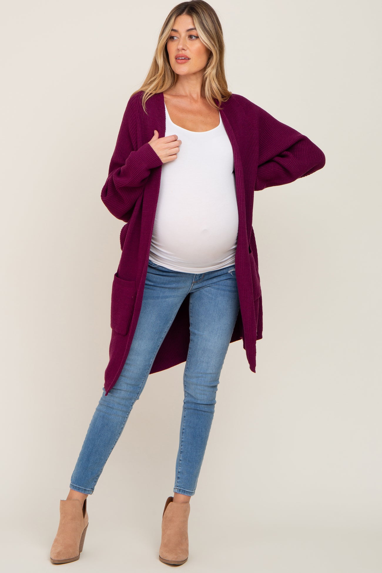 woman wearing a burgundy maternity cardigan over a white top and showing her baby bump, paired with blue skinny jeans