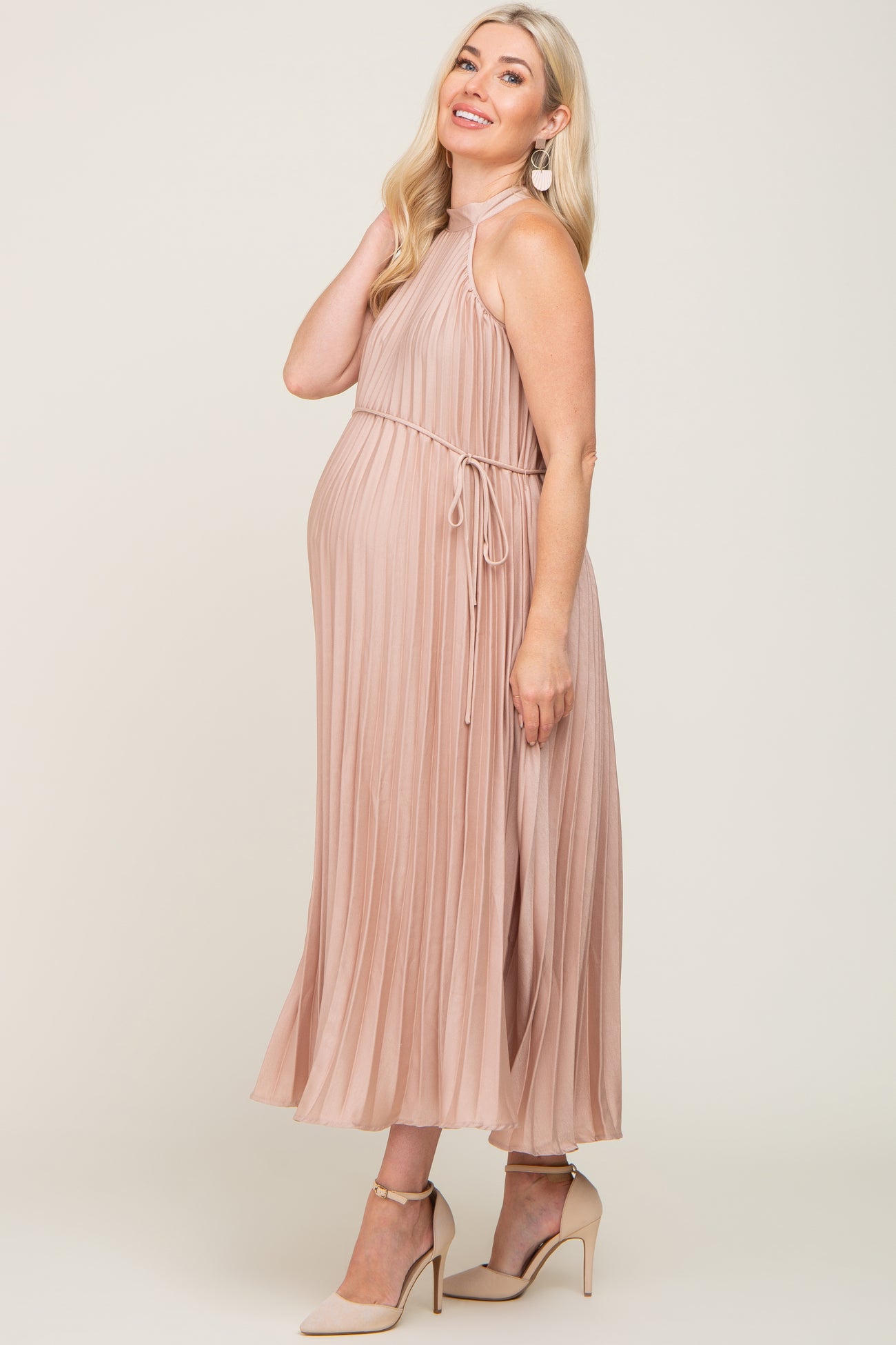 woman wearing a light pink pleated maternity dress with a halter neck and tie waist, showing her baby bump