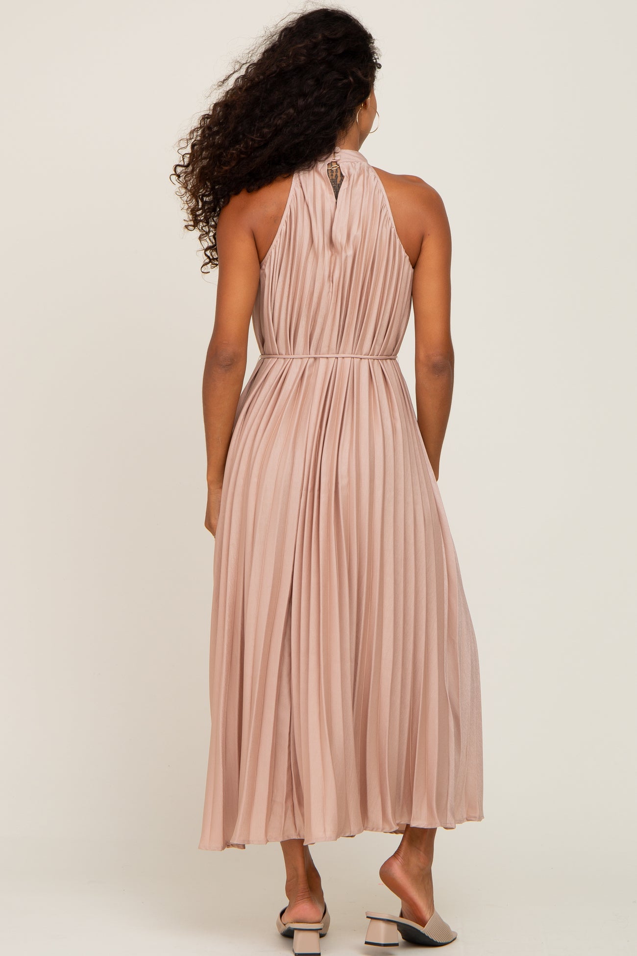 woman wearing a blush pink pleated maxi dress with a halter neck and a cinched waist, viewed from the back