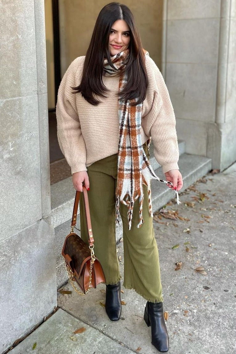 woman wearing a beige oversized sweater, olive green flared pants, and a patterned scarf, holding a brown handbag