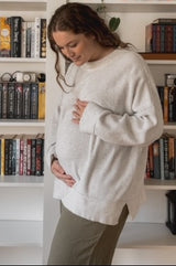 woman wearing a light gray ribbed maternity sweater and olive green pants, gently cradling her baby bump